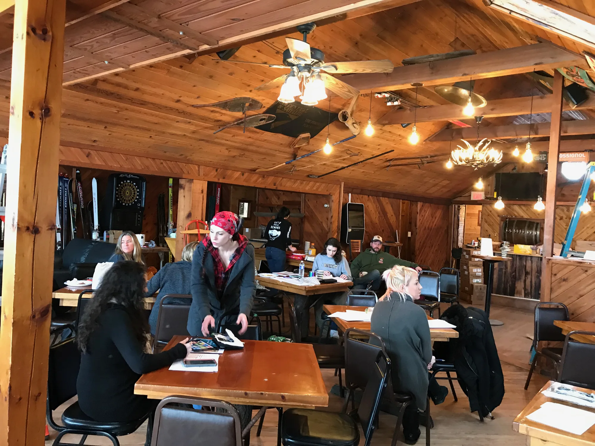 Wood-beamed dining room with people working at tables, an antler chandelier, and skis and snowshoes on the walls and ceiling