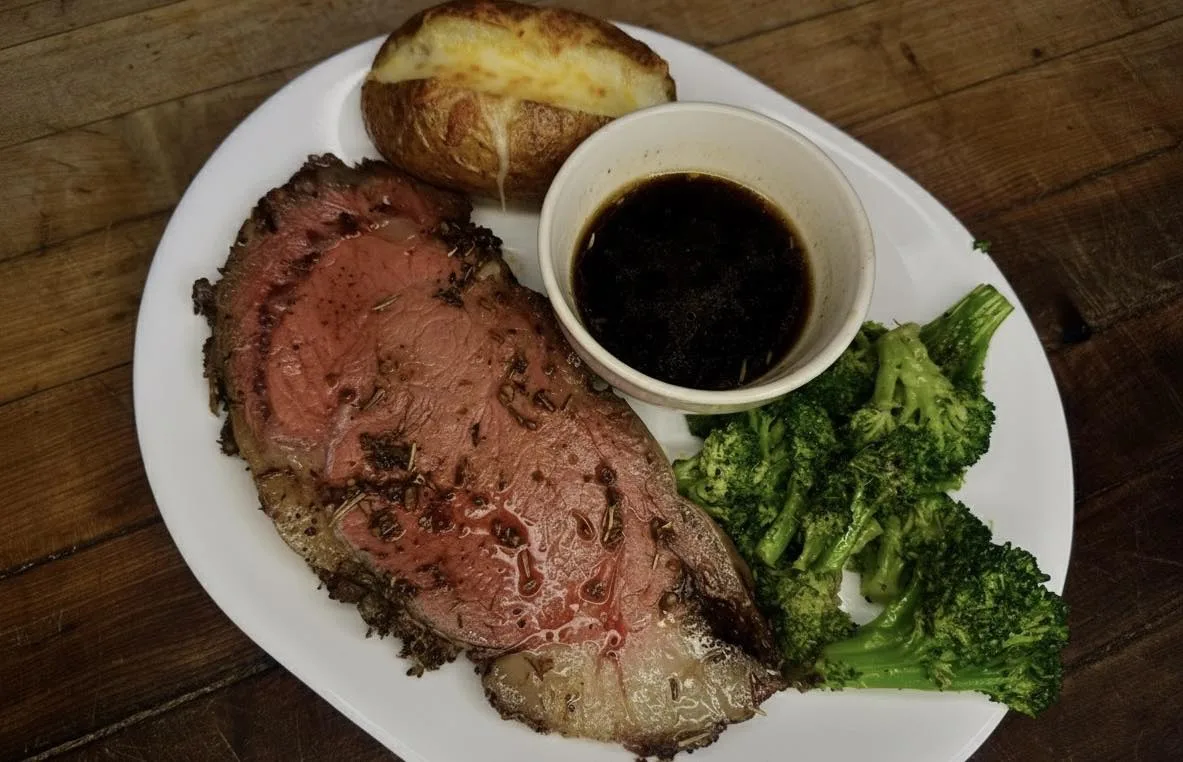 Herb-crusted prime rib with a baked potato, au jus and broccoli on a wood table