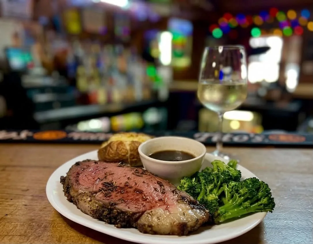 Prime rib with au jus, baked potato and broccoli on the bar, white wine and bar lights behind