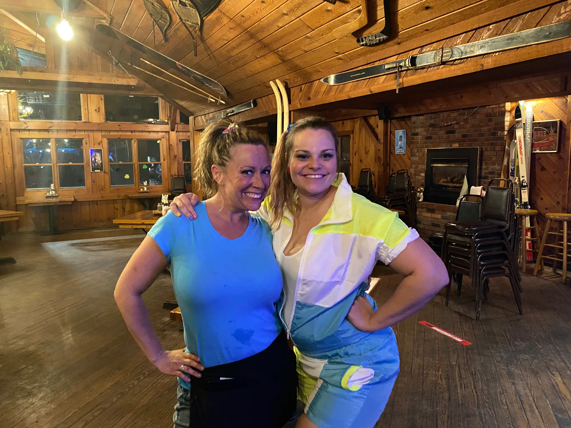 Two smiling servers in the lodge dining room with skis overhead and a brick fireplace